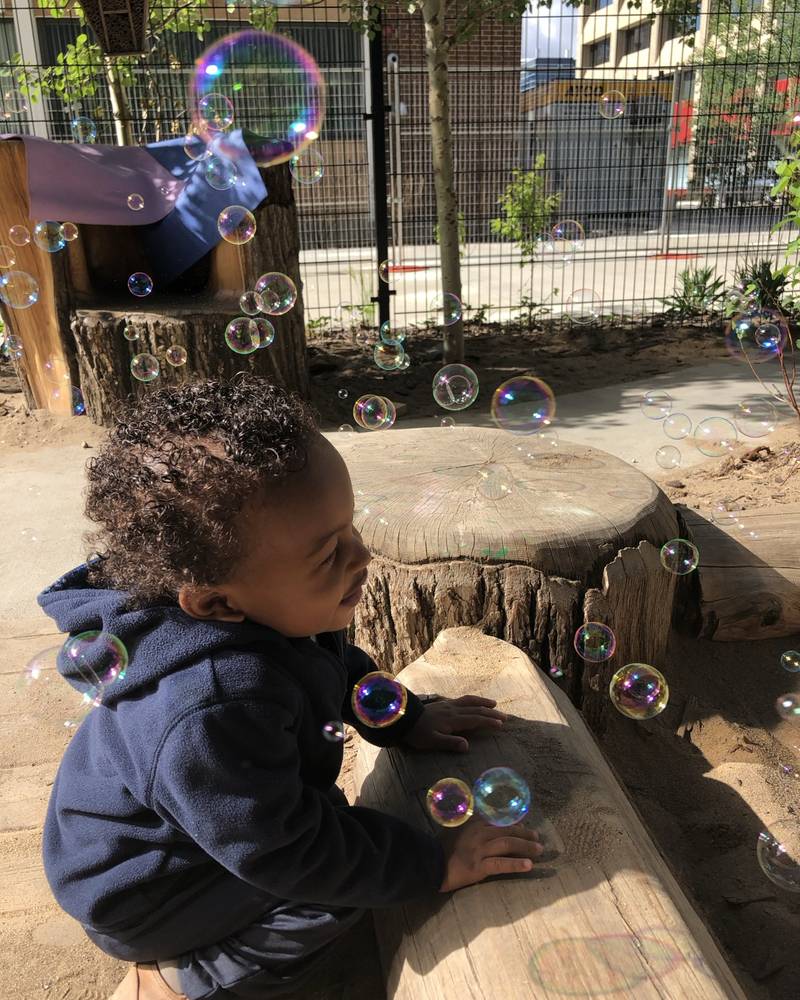 A toddler crouched on a log, reaching for soap bubbles