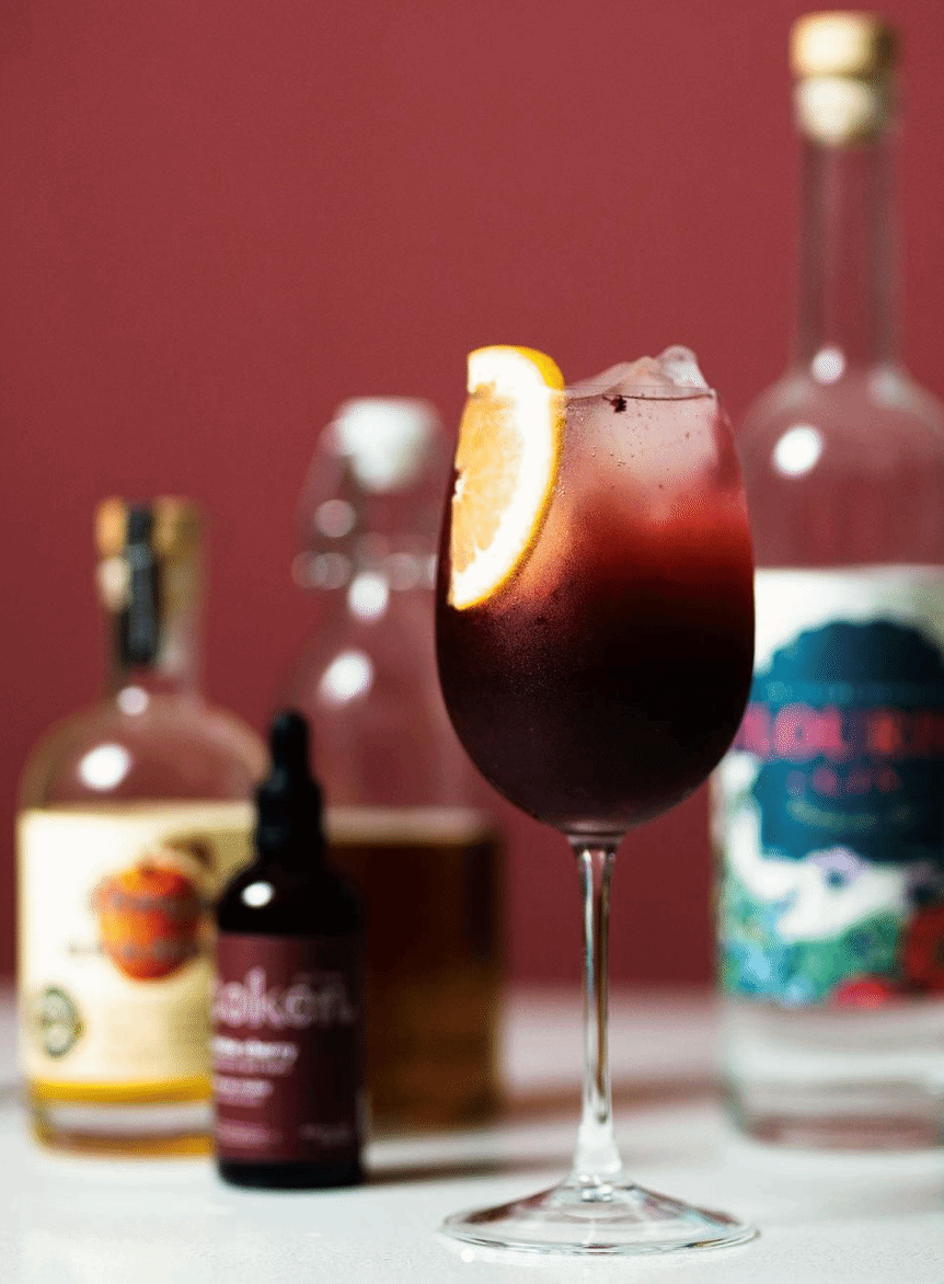 Glass of Sangria, garnished with a orange slice. Background contains bottles of gin, brandy, and cherry bitters.