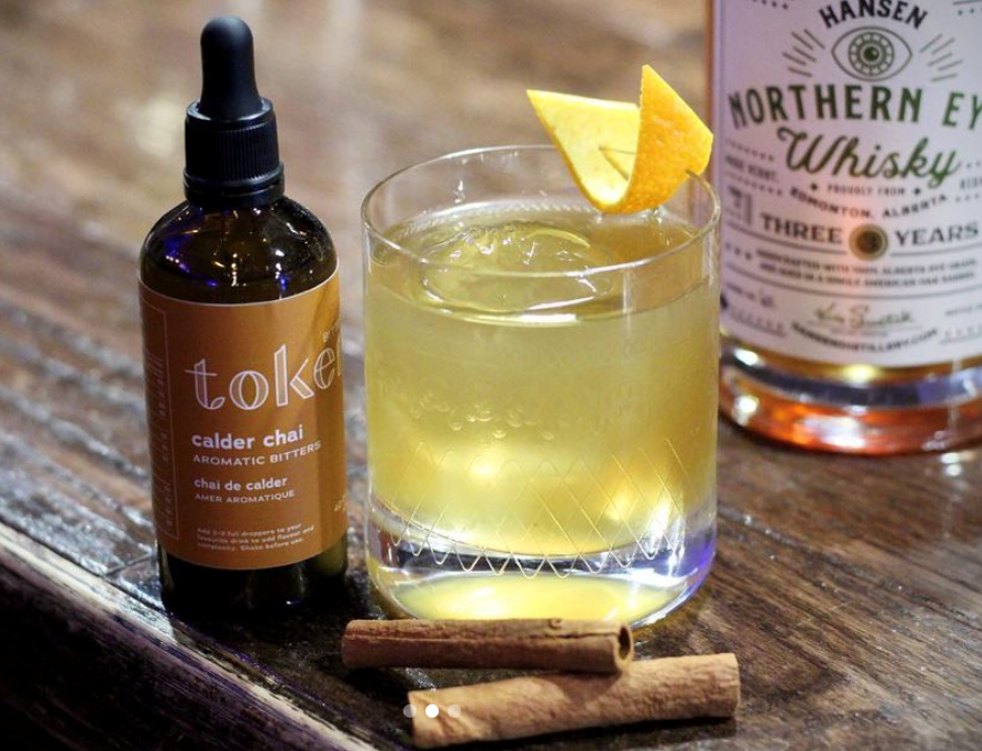 Pale yellow cocktail served over ice in an old-fashioned glass. Served with orange and cinnamon garnish. The cocktail is made with Whiskey and Token Calder Chai Bitters.