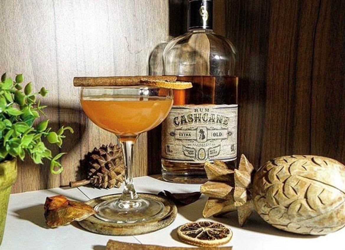 Orange Daiquiri served in a coupe glass, garnished with cinnamon and lemon. Features rum and assorted decorations in the background.