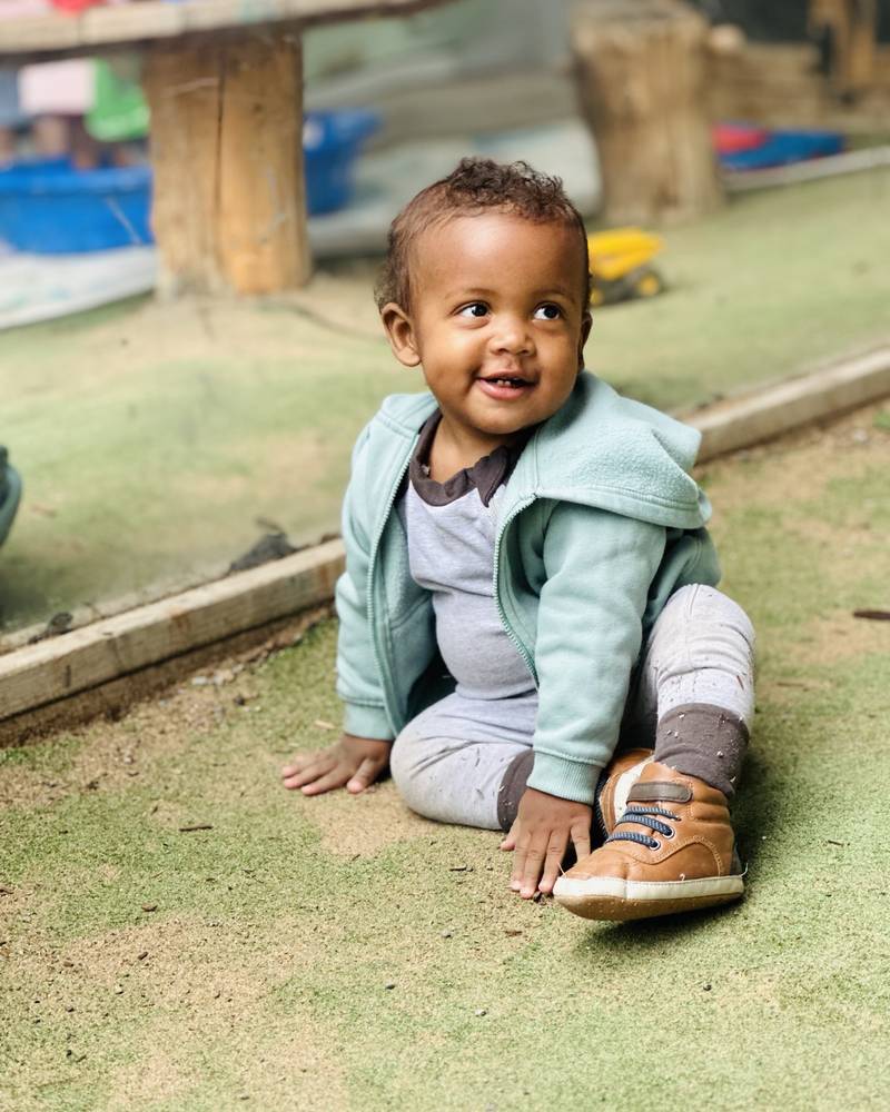 A baby boy in a mint hoodie sitting on the play yard turf, laughing