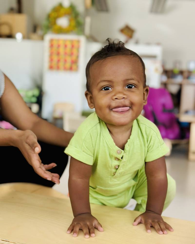 A baby in a lime green romper on a wooden table, beaming at the camera