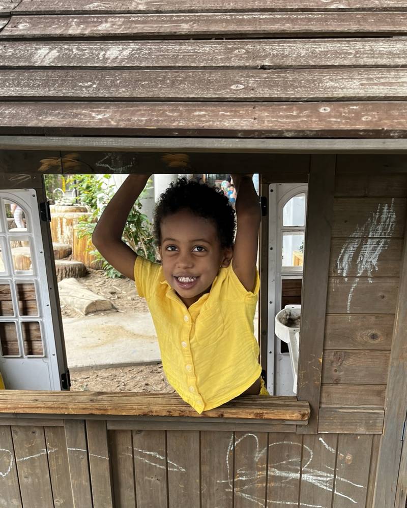 A boy in a yellow shirt hanging from the window of a wooden playhouse