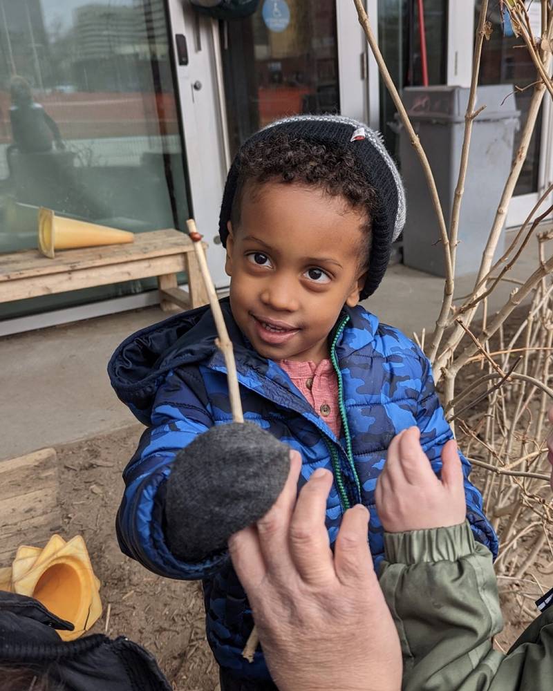 A boy in a blue puffer jacket and toque holding up a stick with a ladybug resting on its tip